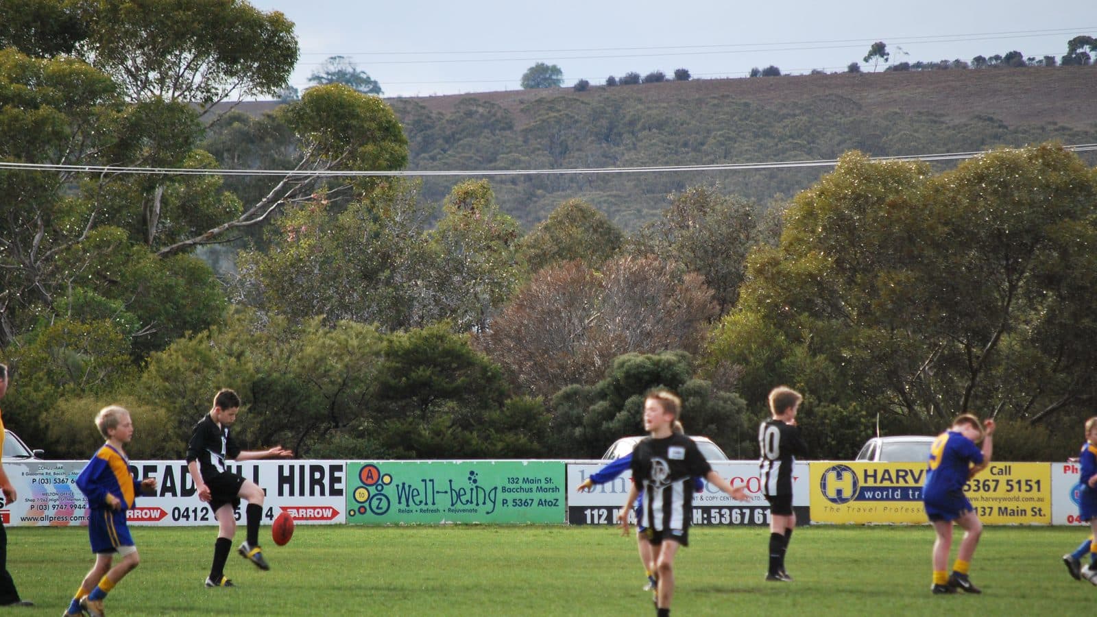 A country football oval during a junior Australian rules match, players in dark and light guernseys scattered across the green grass with trees and low hills behind.
