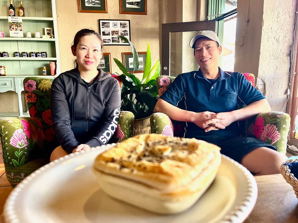 Lisa and Daniel of Chiltern Bakery seated side by side on floral armchairs inside the cafe, with a golden-crusted family-sized pie on a white plate in the foreground and framed black-and-white photographs on the wall behind them.