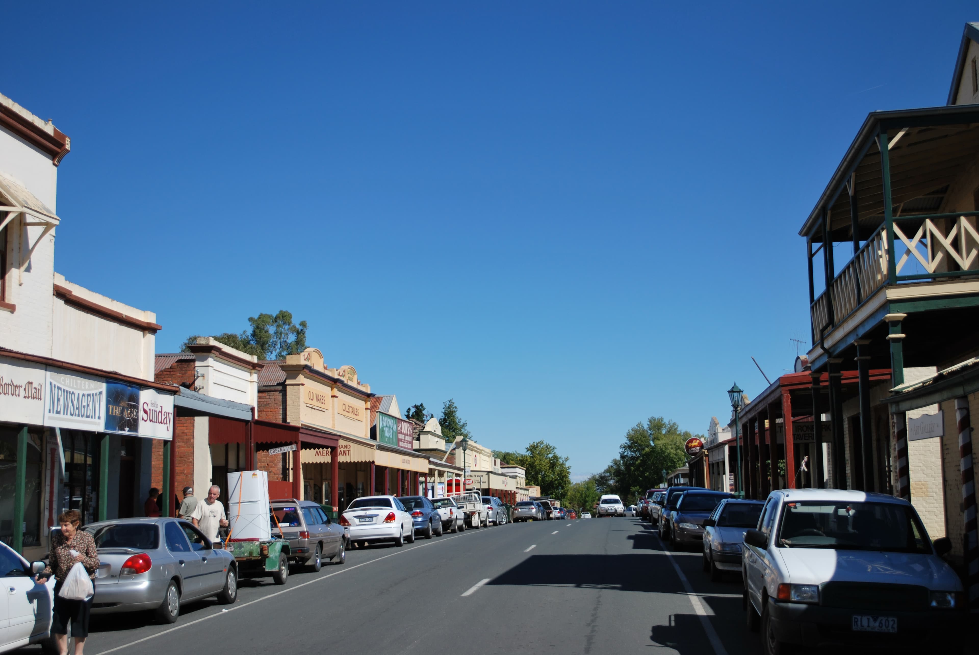 Main Street in Chiltern, Victoria — low historic shopfronts on both sides of a wide country-town road, mature trees, a single car parked at an angle.