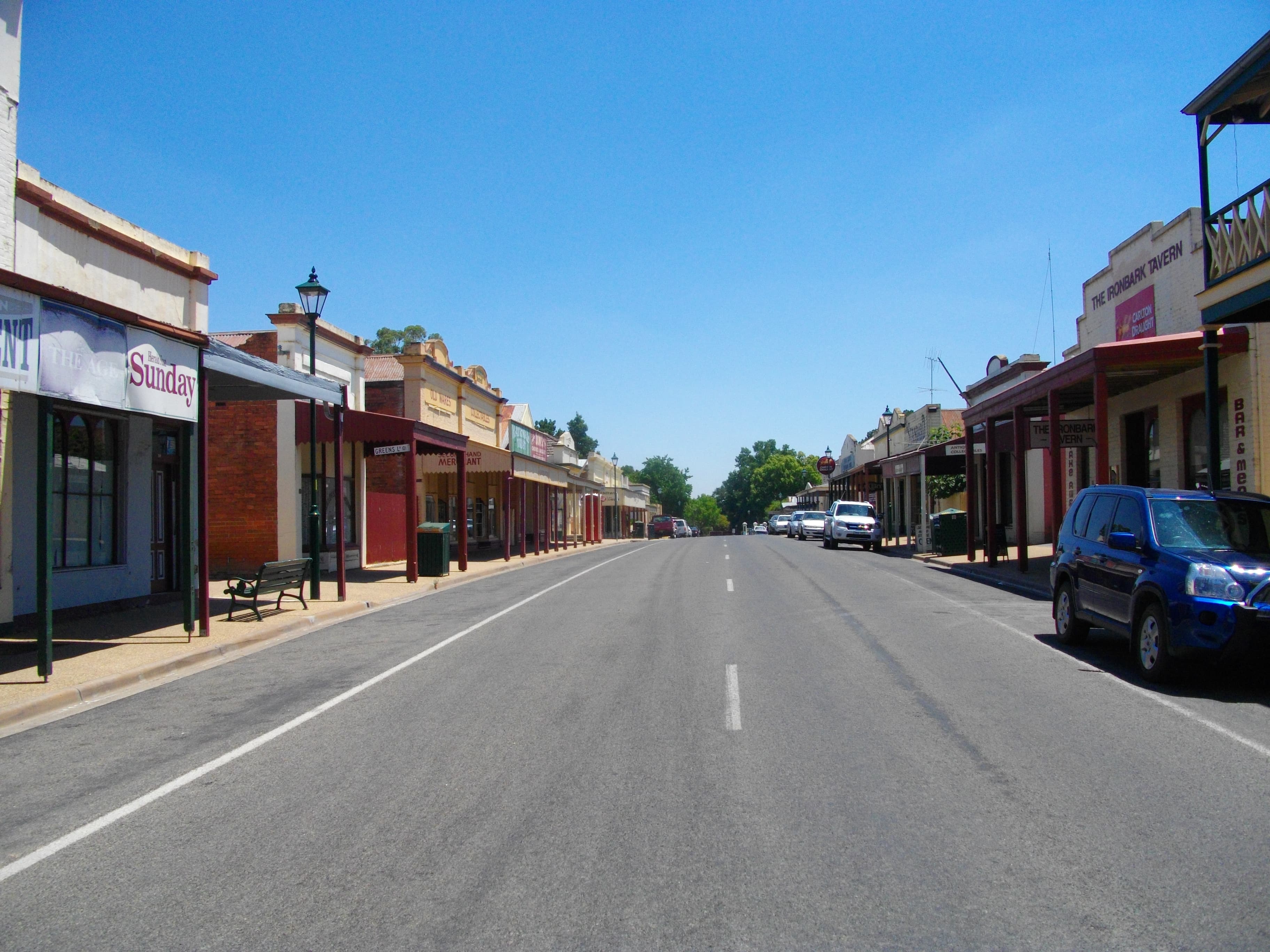 The main street through Chiltern, Victoria, looking past historic shopfronts on both sides — wide road, mature trees, low timber verandahs in winter light.