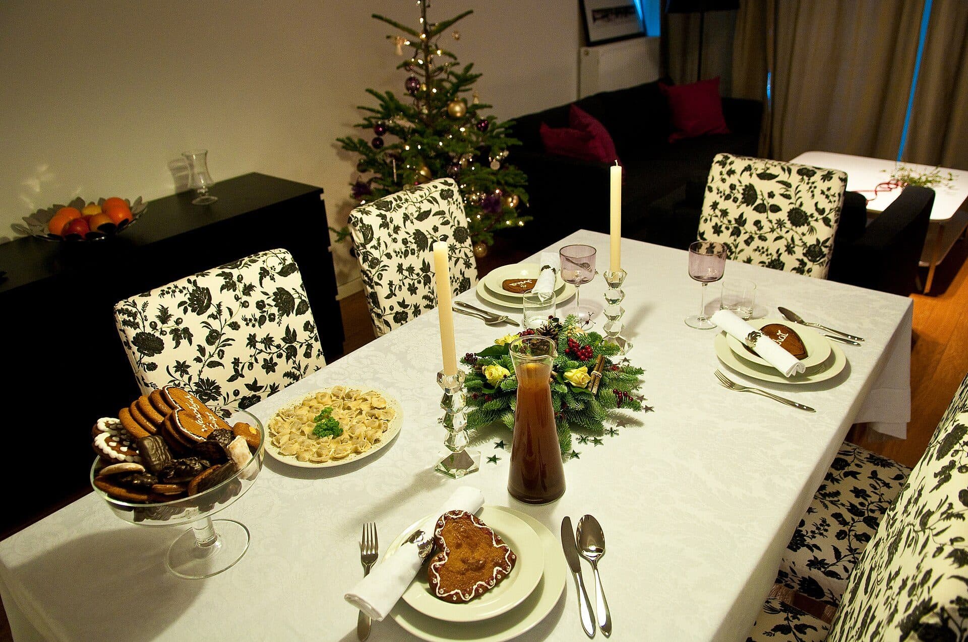 A home dining table set for Christmas dinner in warm evening light: a white damask cloth, two tall lit candles, a pine-and-rose centrepiece, laid place settings, a glass stand of gingerbread, and a decorated, lit Christmas tree glowing behind.