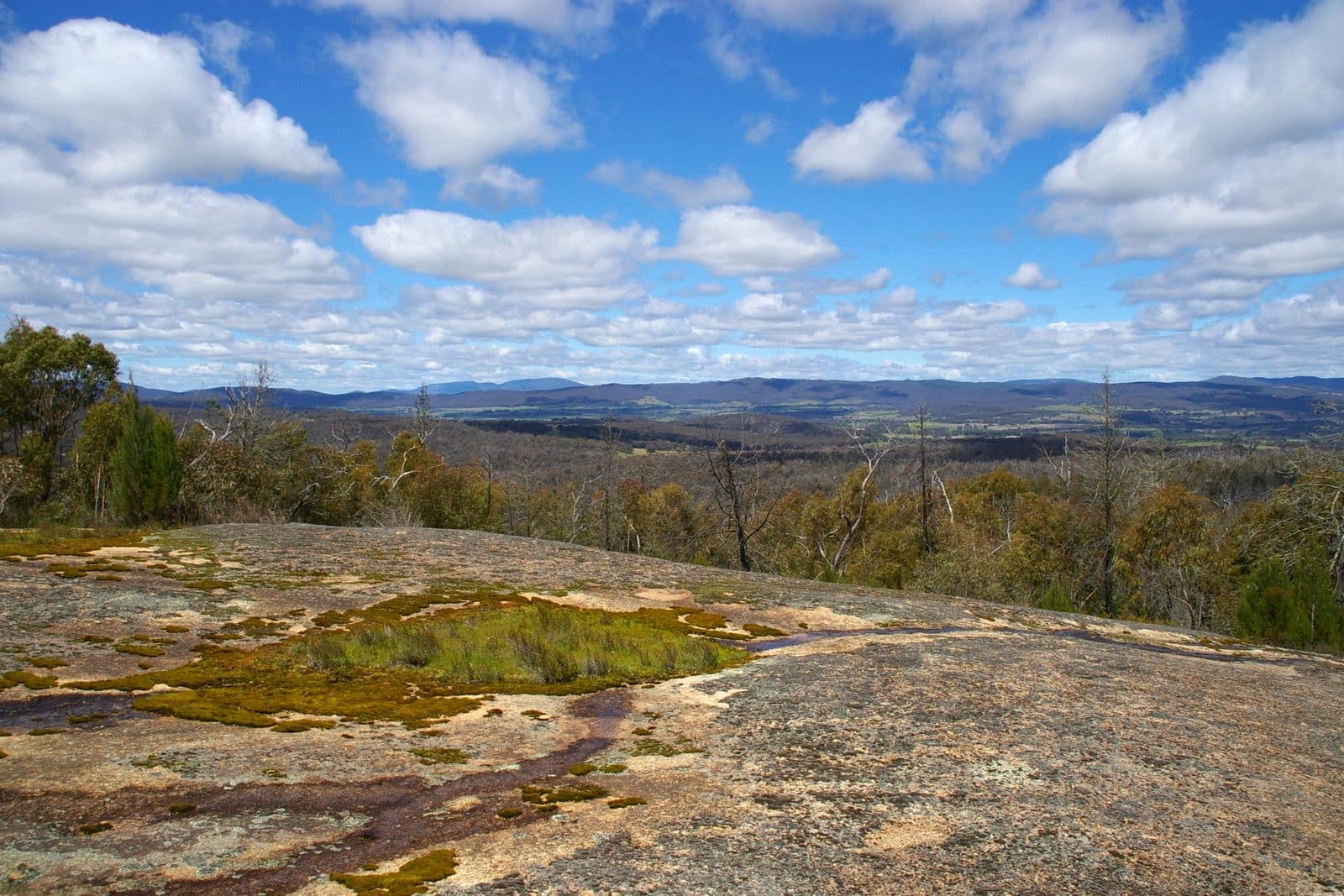 The view from a broad granite outcrop at Mount Pilot summit, looking out over wooded ridges and farmland to distant blue hills under a sky of scattered cloud.