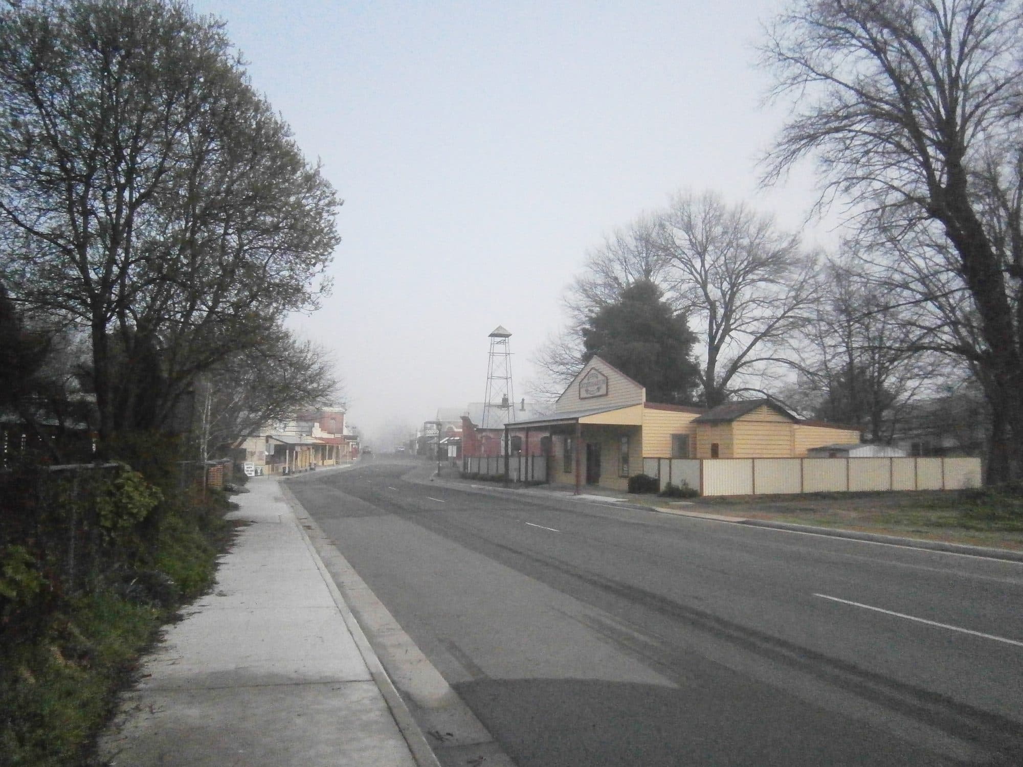 Conness Street in Chiltern, Victoria, looking west โ wide road, low historic shopfronts on both sides, mature trees, a quiet autumn morning.