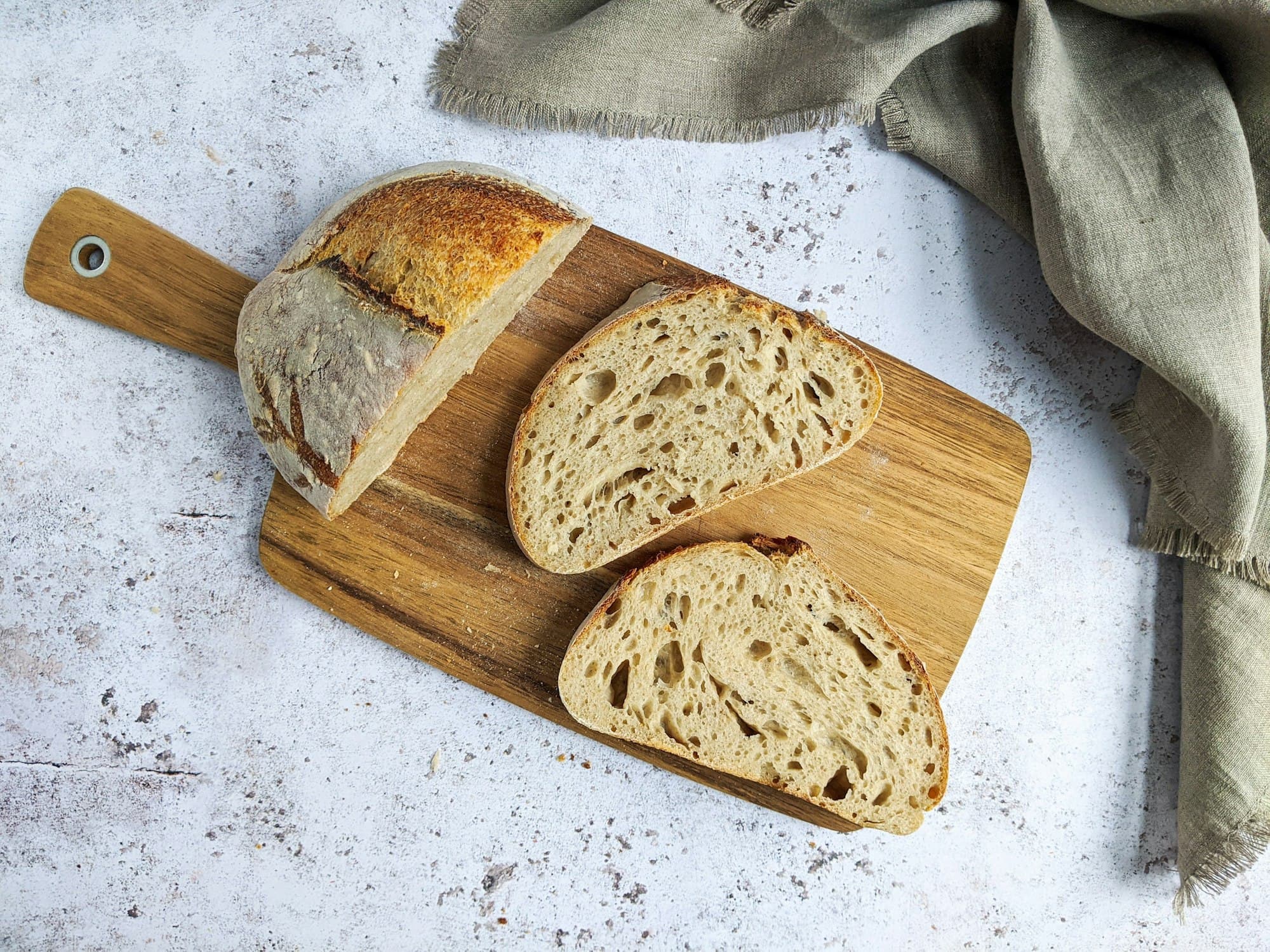 A whole wholegrain loaf resting on a wooden cutting board, uncut, with a dark scored crust and a dusting of flour across the top.