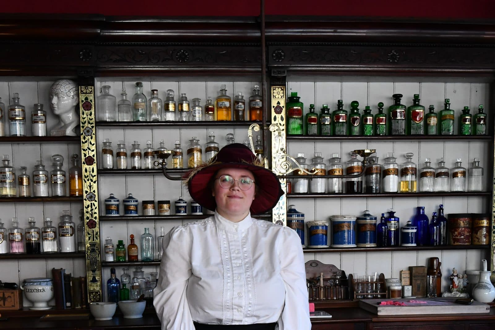 The inside of a preserved chemist shop, shelves of labelled glass bottles running the width of the wall behind a timber counter, with a person in period dress standing behind it.