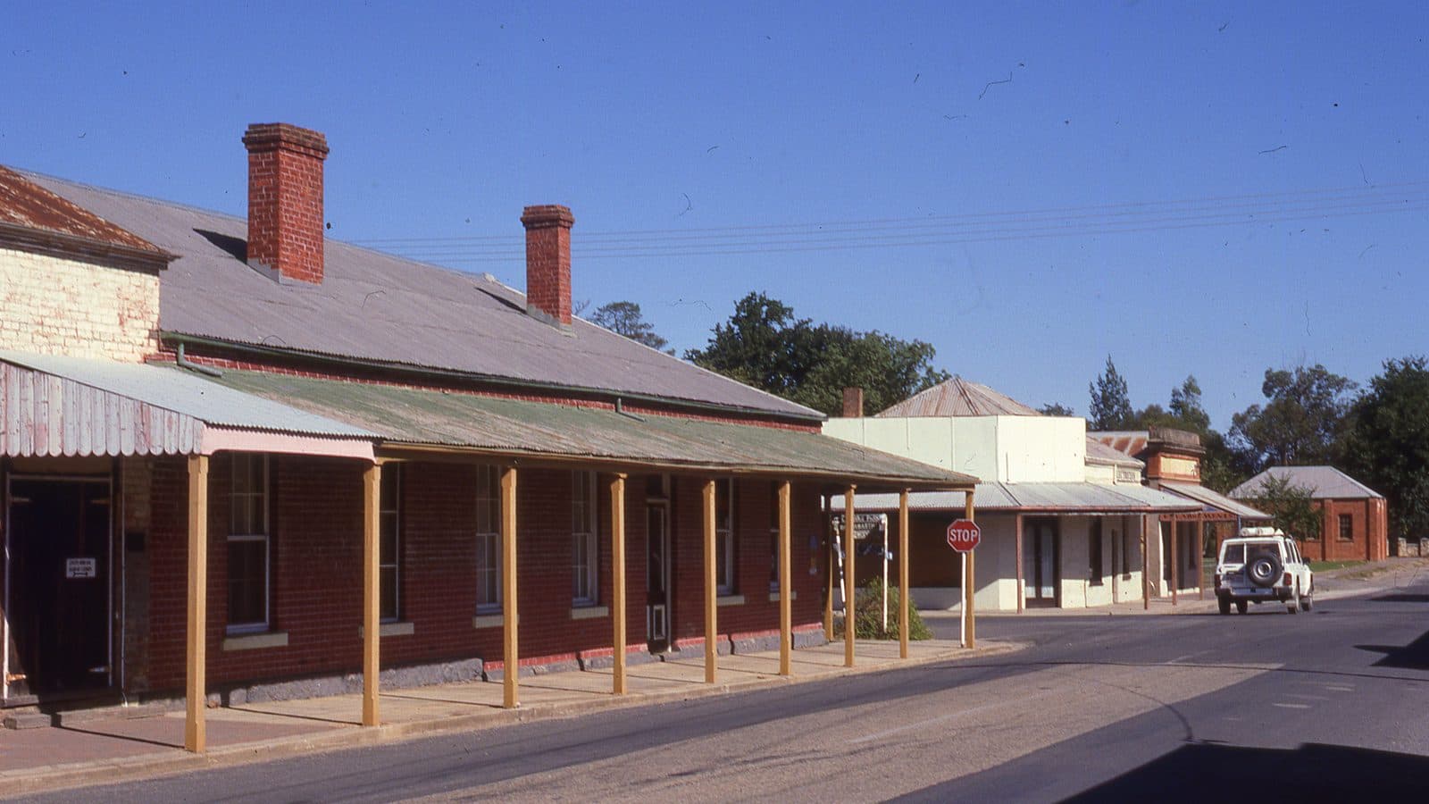 A quiet country street with a long single storey red brick building under a timber post verandah, two brick chimneys along its roof, and older shopfronts further down past a stop sign.