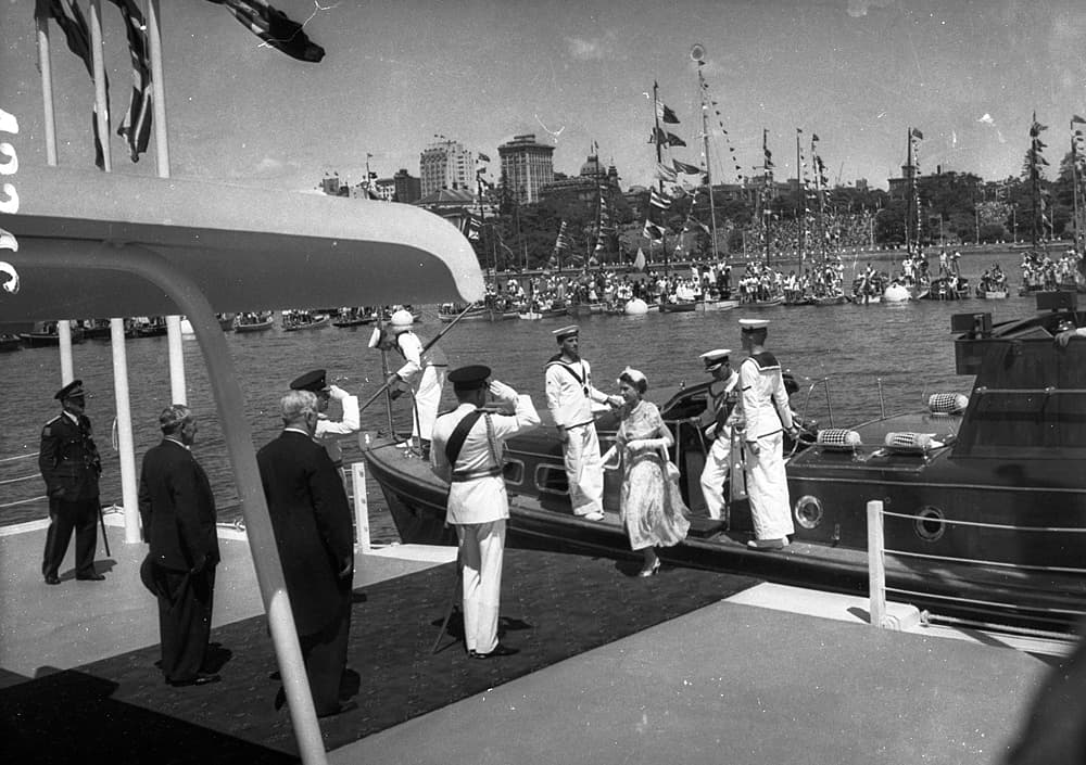 Queen Elizabeth II in a pale coat and hat steps ashore at Farm Cove, Sydney, during the 1954 royal tour, with officials and a small crowd gathered behind her.