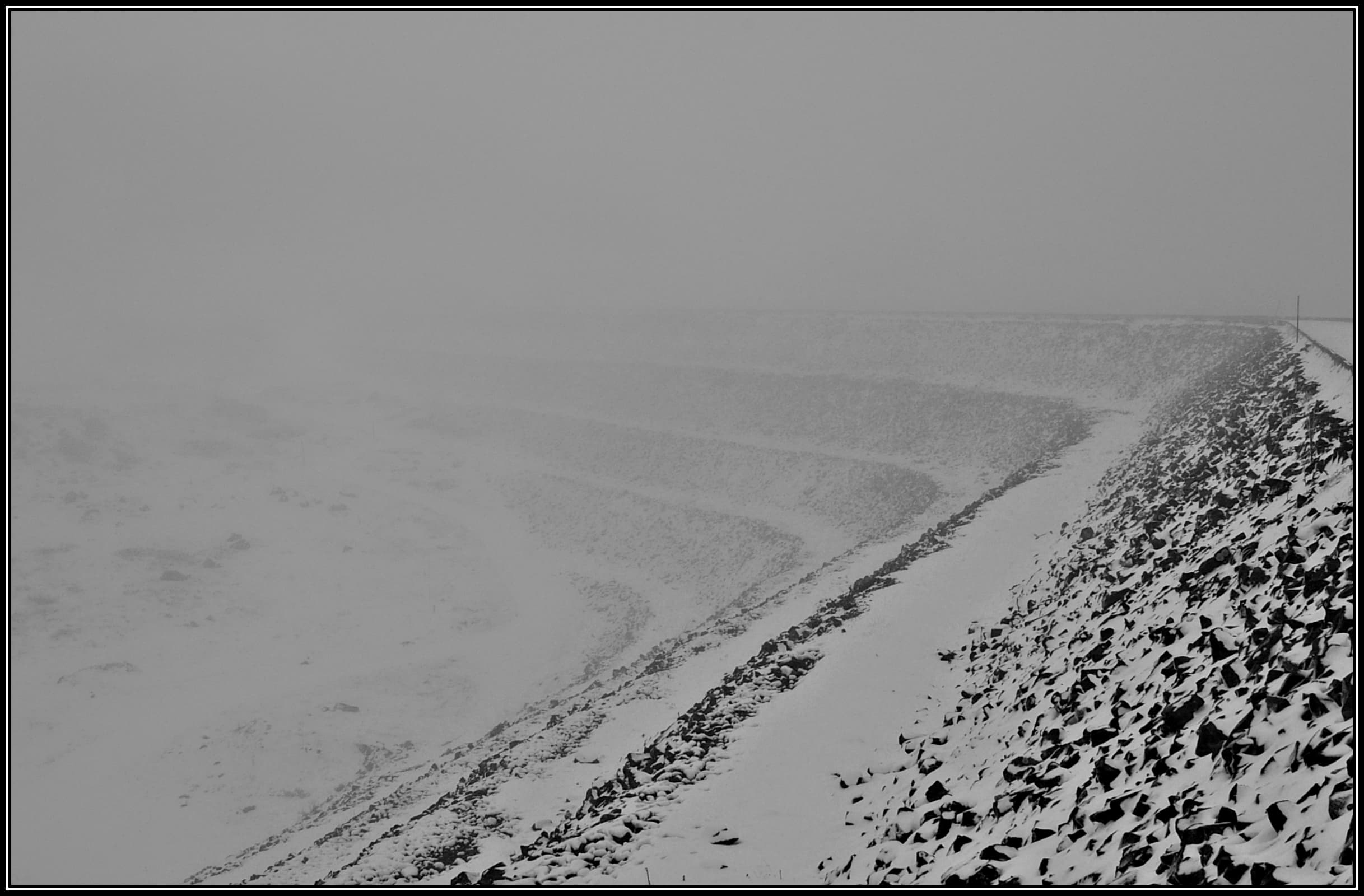 Snow and mist closing over the Rocky Valley Storage Dam in the Victorian high country โ a pale, near-monochrome winter landscape with the far shore lost in cloud.